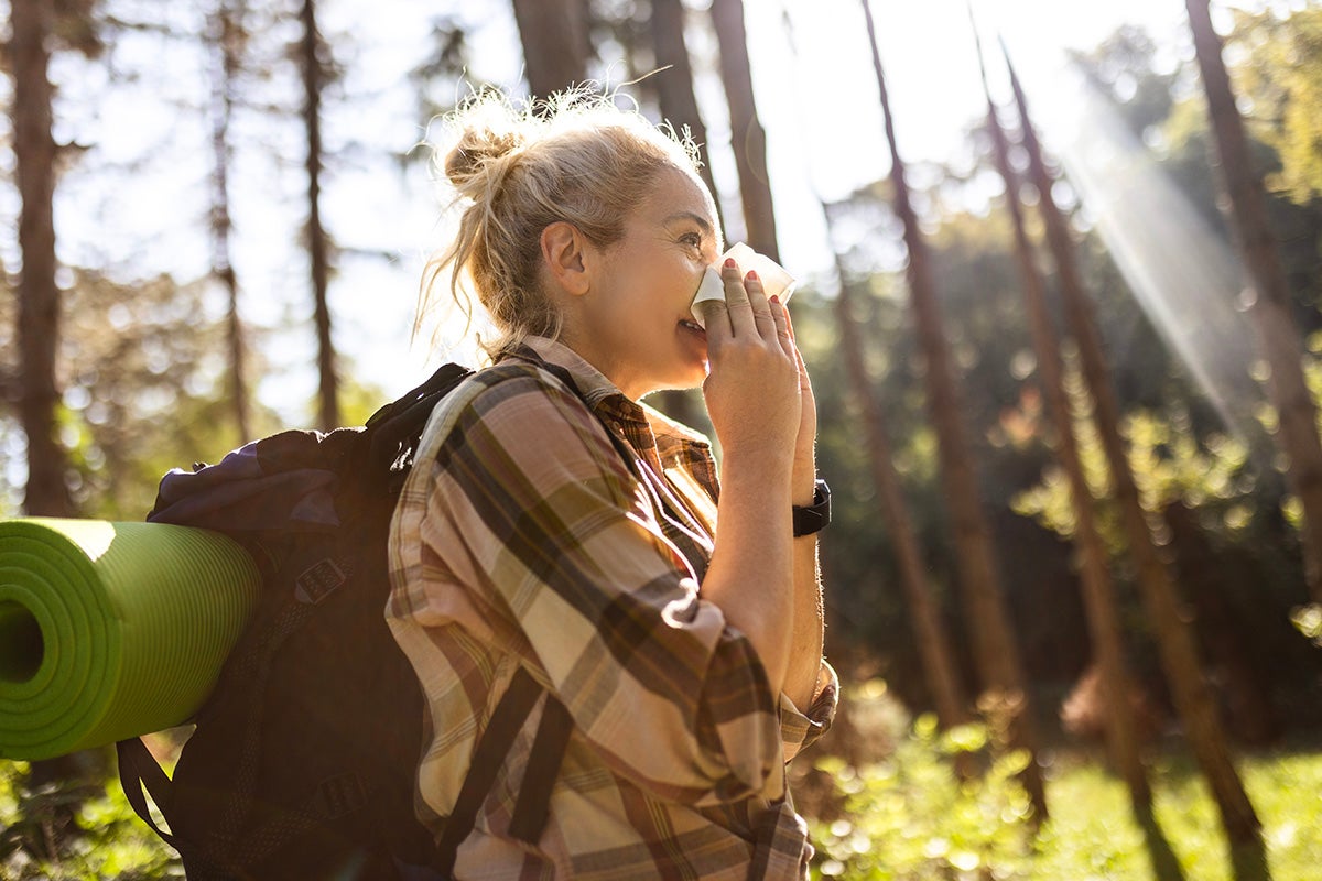 Woman with a backpack blowing her nose with a tissue while hiking in a sunny forest, suggesting seasonal allergies outdoors.