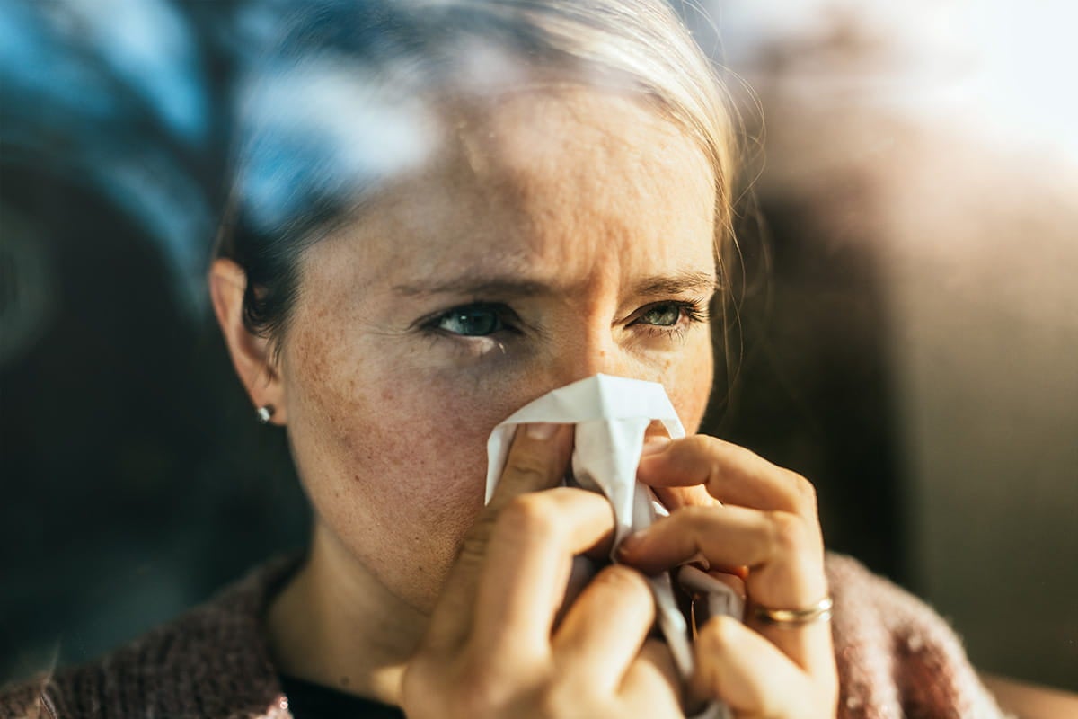 Woman blowing her nose with a tissue, showing cold or allergy symptoms indoors.