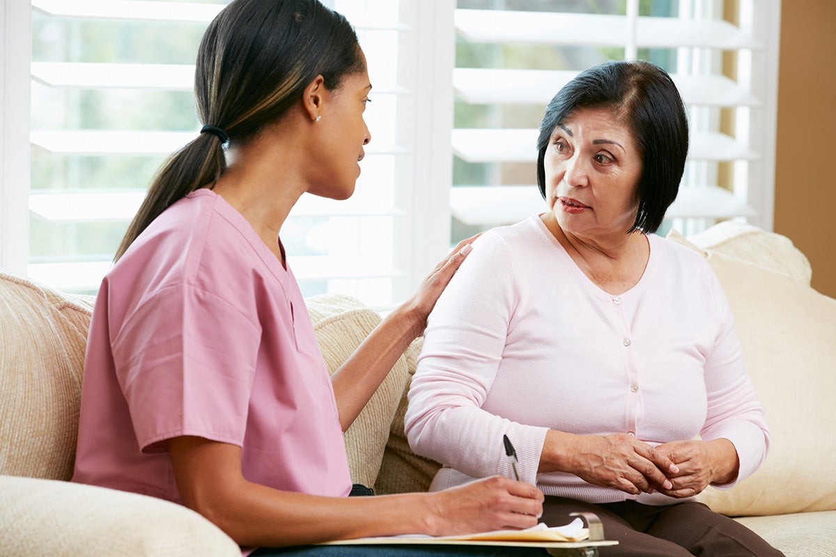 A caregiver in scrubs gently speaking with an older woman on a couch, offering support while taking notes in a home setting.