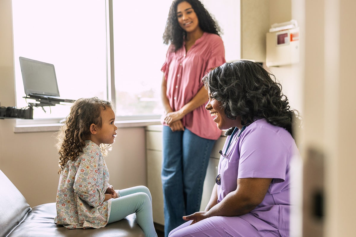A pediatric nurse smiles while speaking with a young child seated on an exam table.