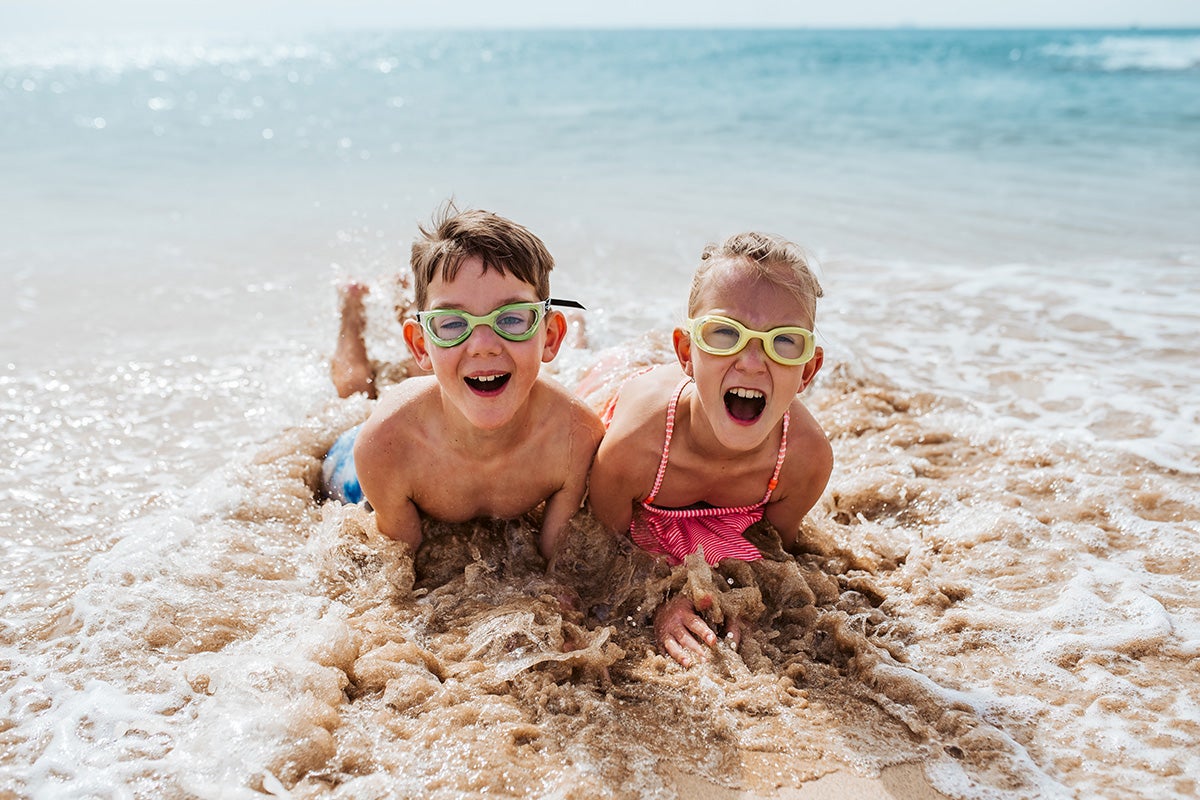 Two children wearing goggles play and splash in ocean waves at the beach, promoting summer activity and outdoor fun.