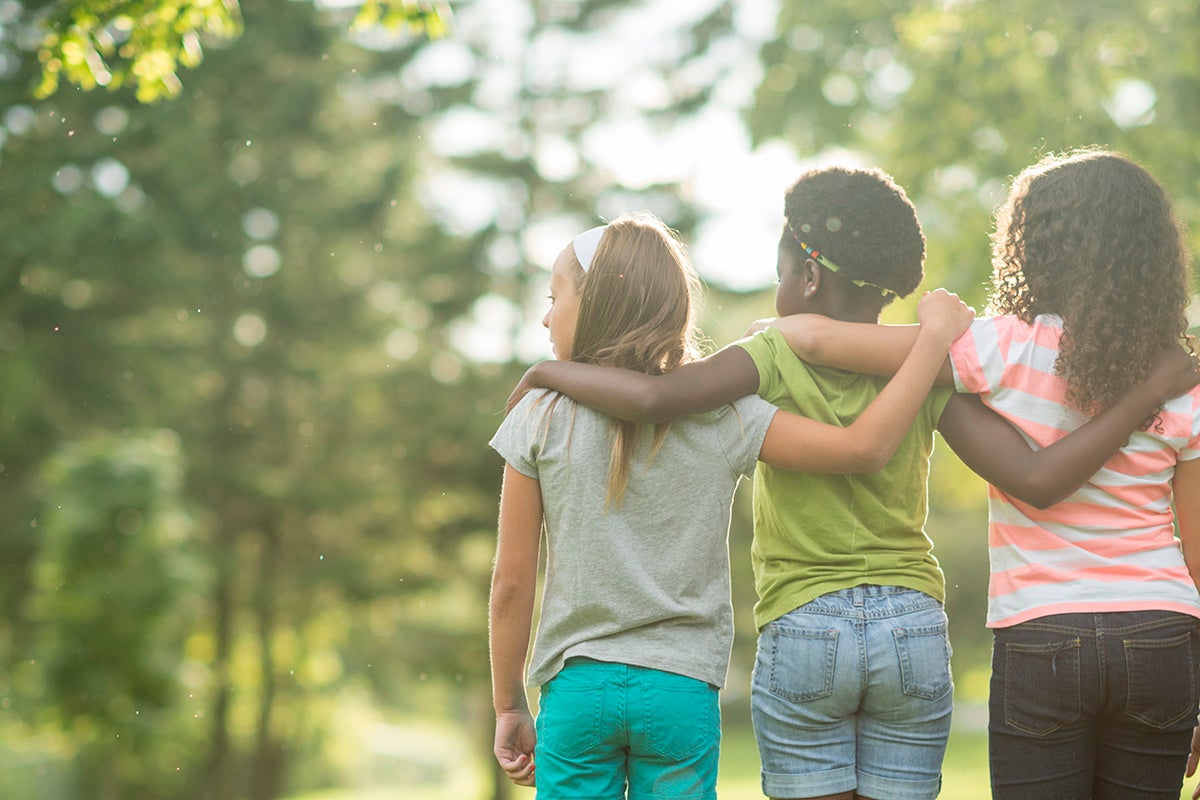 Three children walking outdoors with arms around each other in a sunlit park.