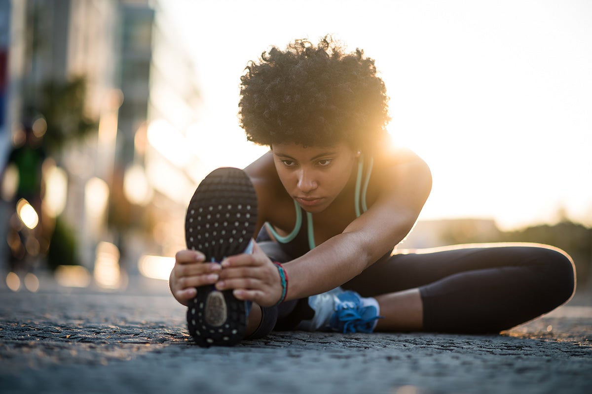 A focused woman stretching her leg on a city street at sunrise, performing a seated hamstring stretch in athletic wear.