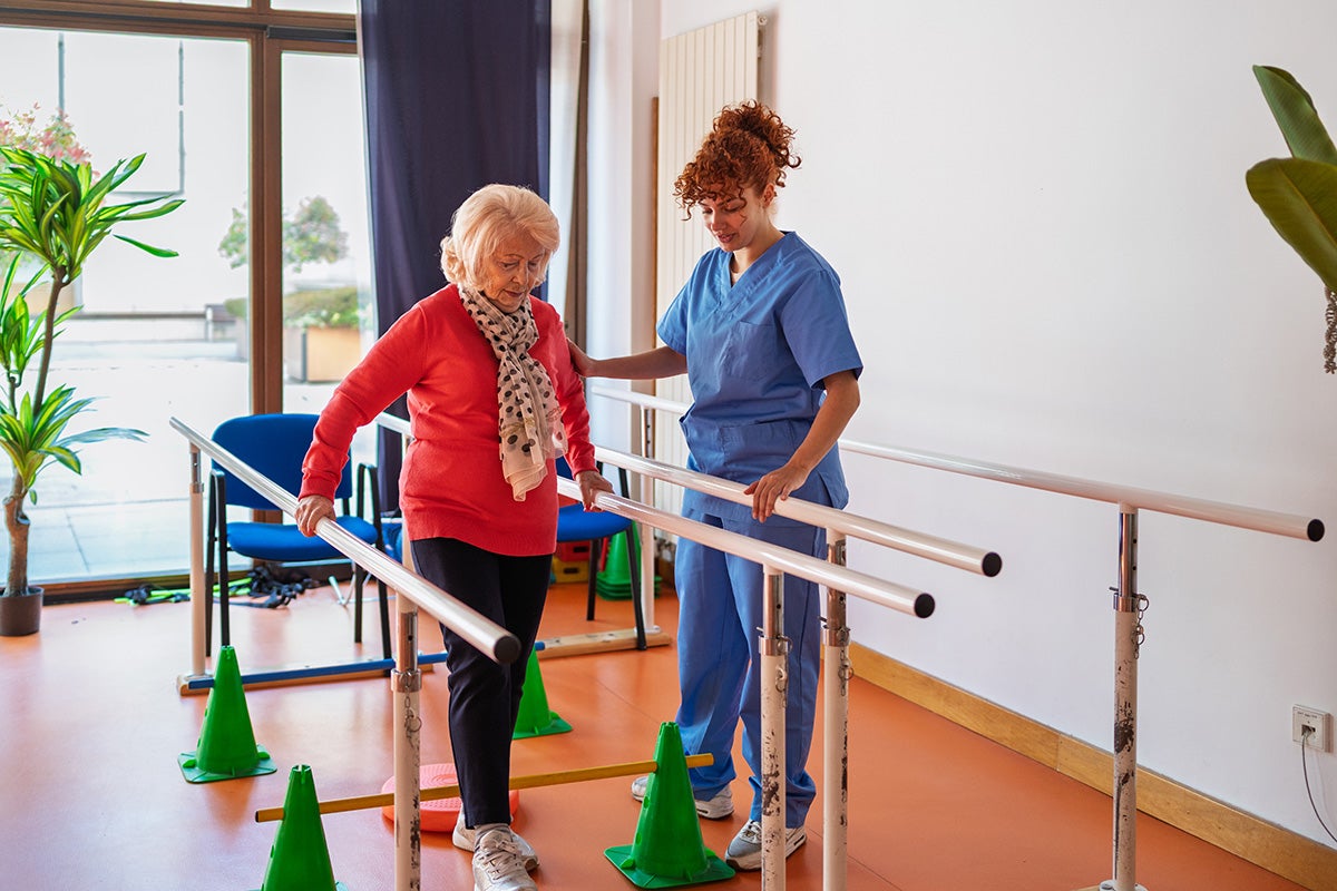 Patient practices walking with parallel bars while a clinician provides support during rehabilitation.