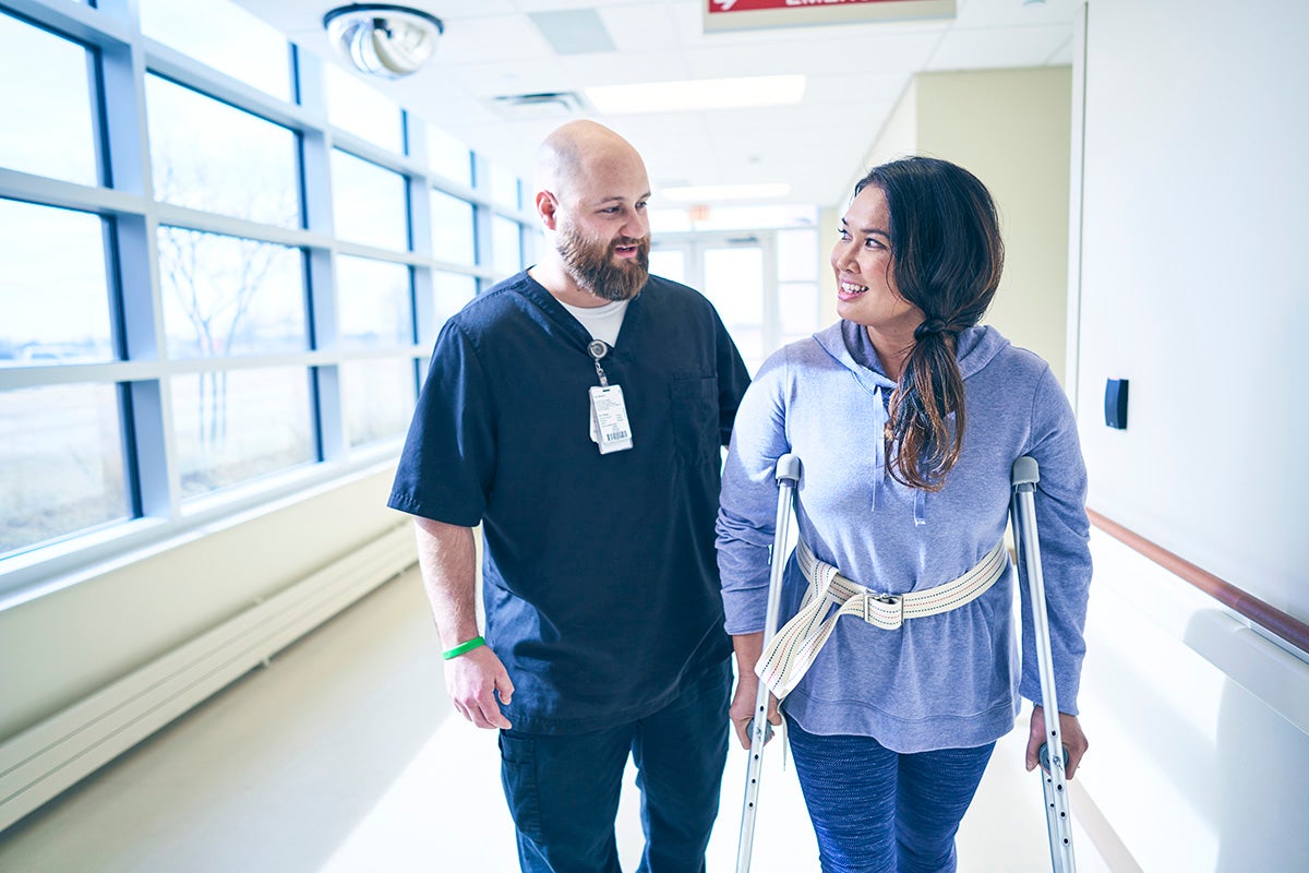 Ascension caregiver supports a patient using crutches while walking in a hospital hallway.