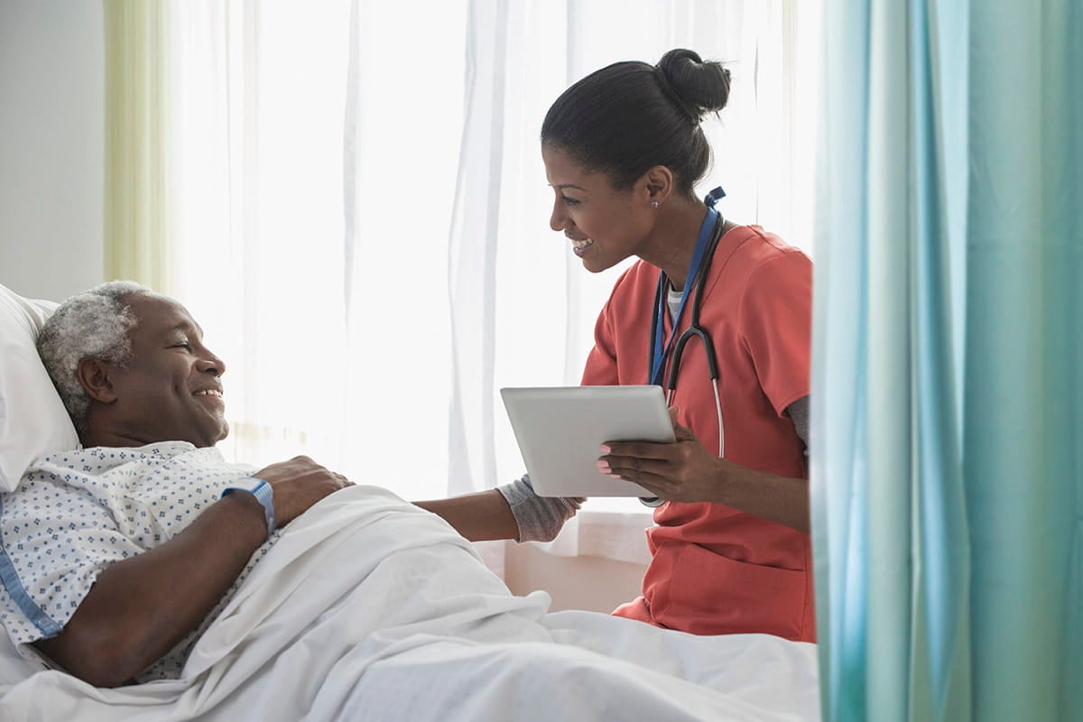 Nurse holding a tablet while speaking with a patient in a hospital bed.