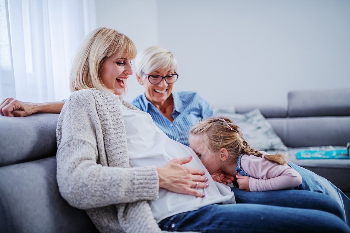 Pregnant woman sitting on a couch as a child leans in to kiss her belly, with another adult smiling nearby.