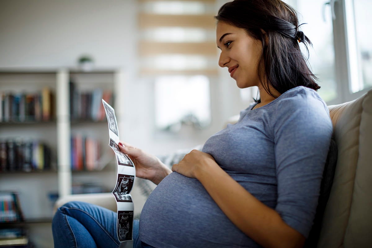 Pregnant woman sitting on a couch holding and looking at ultrasound images.