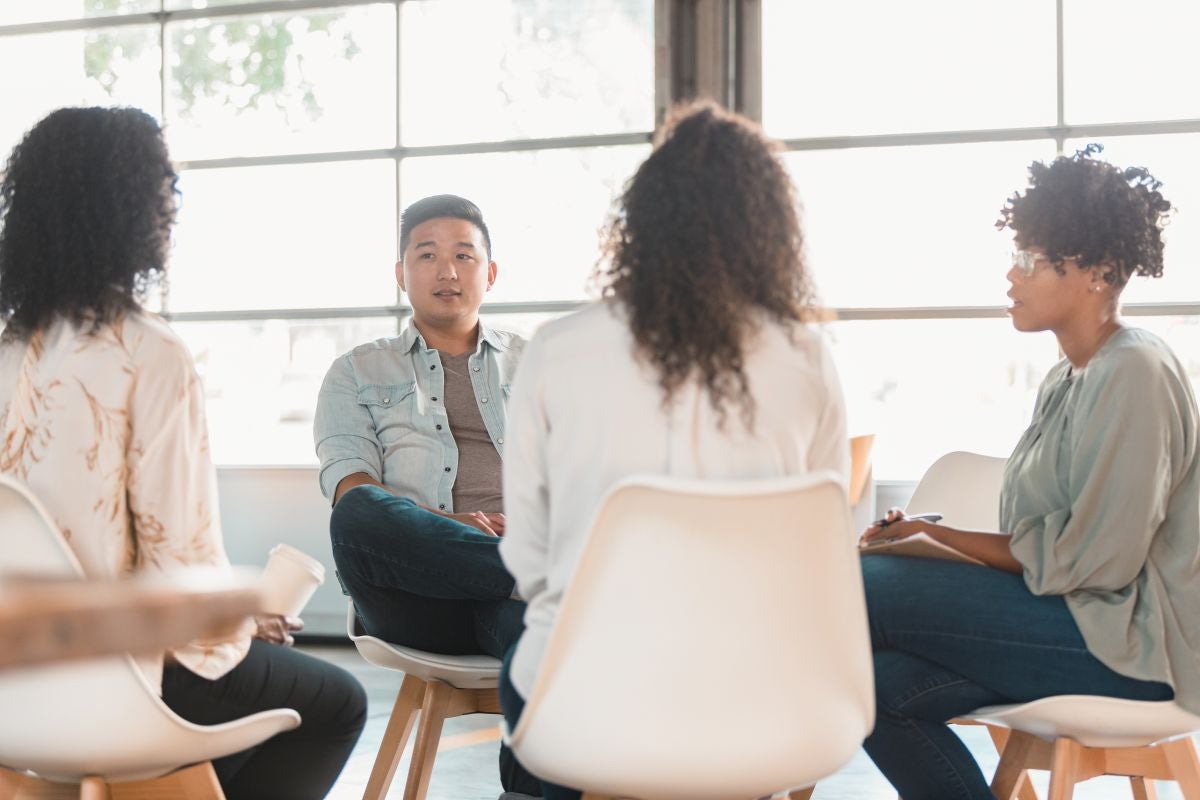 Four adults sitting in a circle having a group discussion in a bright room with large windows.