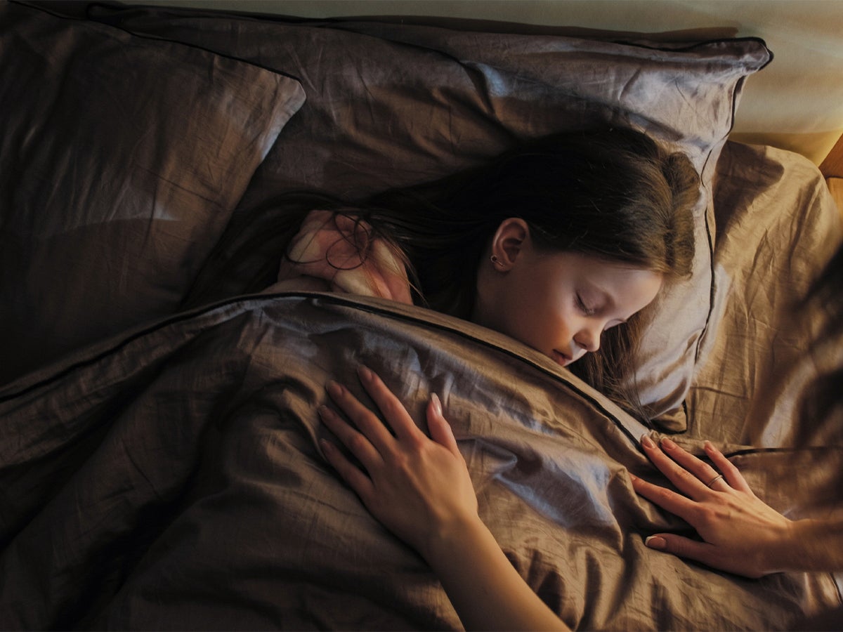 A young child sleeps peacefully in bed while an adult gently tucks in the blanket, illustrating the importance of healthy bedtime routines before the start of the school year.