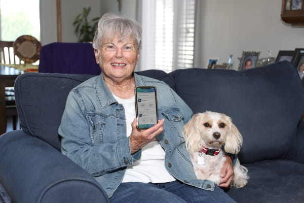 Jean Beck holds up mobile phone with knee mobility-tracking app on screen while sitting on couch with her dog, Belle