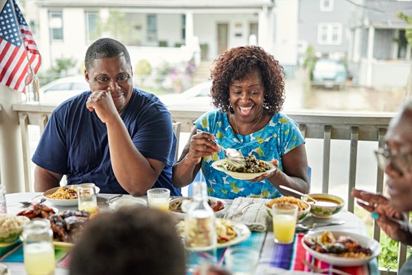 An American family is eating a meal during a Fourth of July holiday celebration. An American Flag is waving in the background.