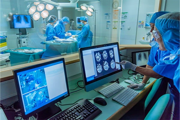 Medical staff in surgical attire monitor a live operation from a control room, viewing patient scans and operating room video on computer screens through a glass window.