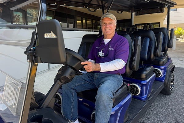 Tommy photo: Ascension St. Vincent’s volunteer Tommy Mouro sits behind the wheel of a large, blue multi-passenger electric shuttle.