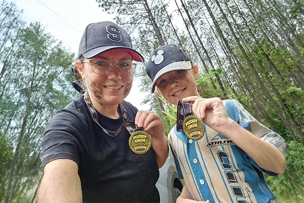 Marie and her daughter smile and hold up their Muddy Dash 5K finisher medals while standing on a wooded trail after completing the race.