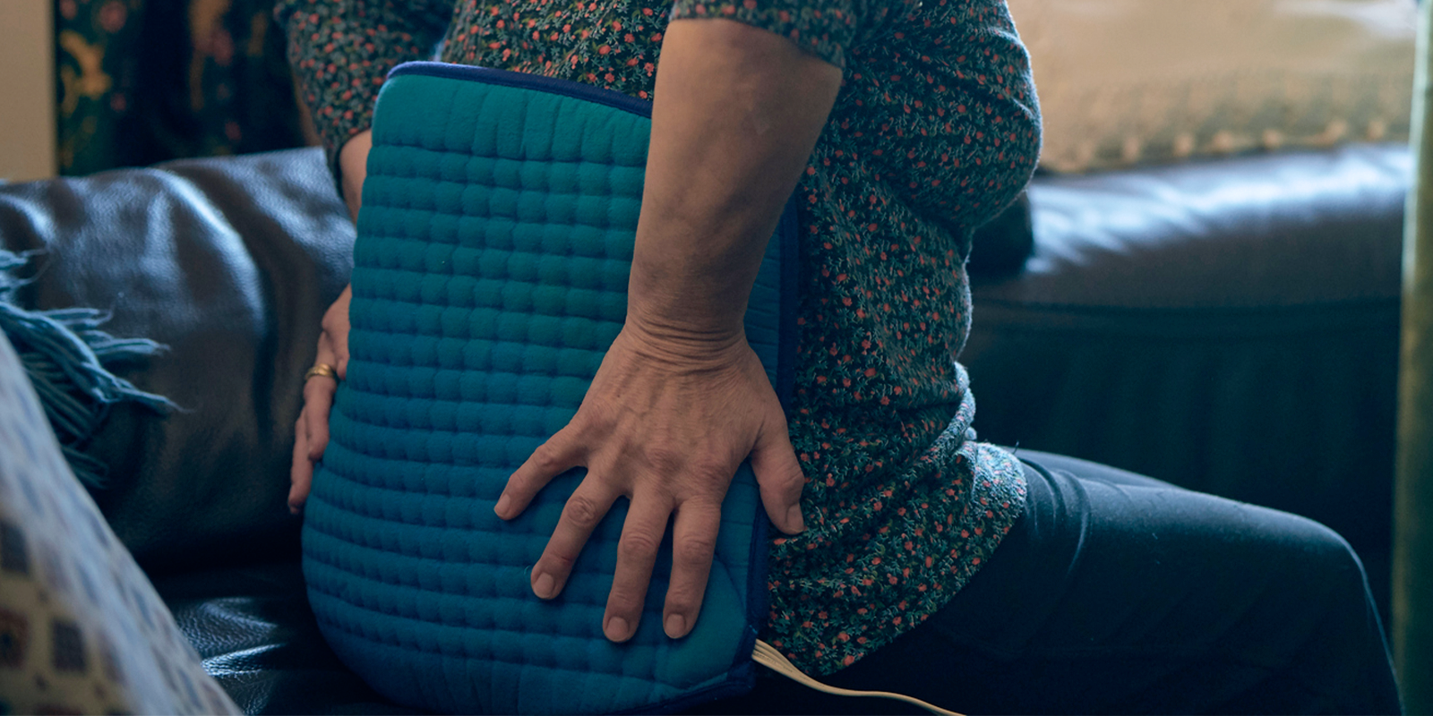 A person holding a blue heating pad against their lower back while seated on a couch.