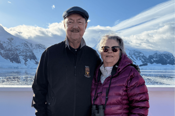 An older couple stands together on a boat deck with snow-covered mountains and icy water in the background