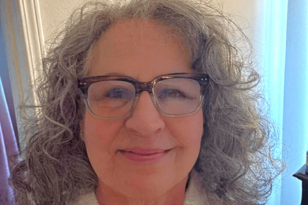 Close-up portrait of a smiling woman with curly gray hair and glasses indoors.