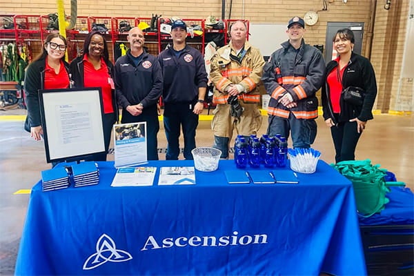 Ascension staff and firefighters standing behind an information table at a fire station during a community outreach event.