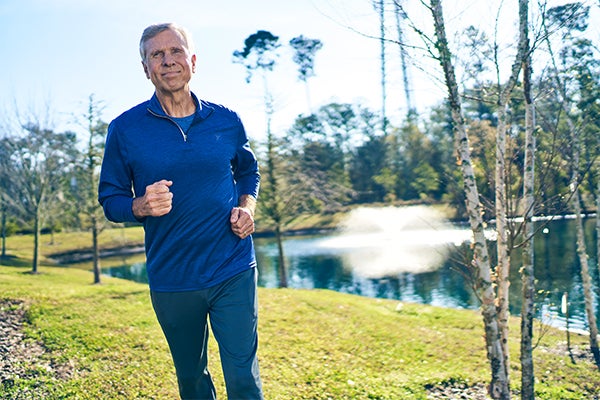 Older adult man jogging along a grassy path beside a pond with a fountain, trees in the background on a sunny day.