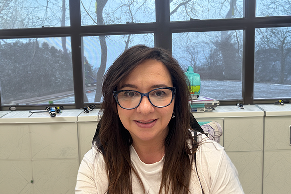 Woman with long brown hair and glasses smiles at the camera indoors, with a window showing leafless trees and a cloudy sky in the background.  