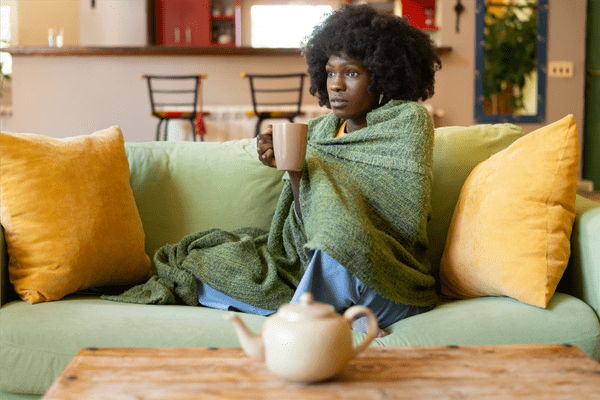 Person sitting on a light green couch wrapped in a green blanket, holding a mug with both hands; a teapot sits on a wooden coffee table in the foreground, and a kitchen is visible in the background.