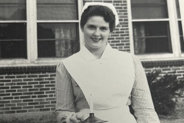 Black-and-white photo of a young nurse standing outdoors in front of a brick building, smiling while wearing a traditional nurse’s uniform and cap and holding a small candle holder