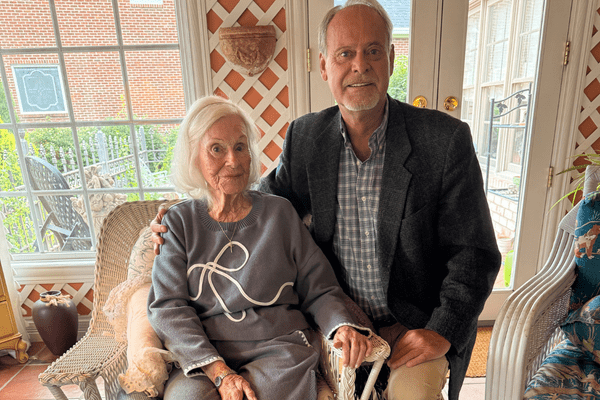 Tim Kempf kneels beside his mother, Edwina Kempf, as she sits in a chair in a bright sunroom.