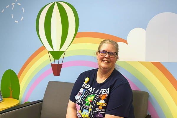 Cristi Hailey, Child Life Champion at Via Christi St. Francis in Wichita, Kansas, smiles while seated on a couch in a brightly decorated pediatric unit, with a rainbow mural and hot air balloon behind her, reflecting a welcoming and comforting environment for children and families.