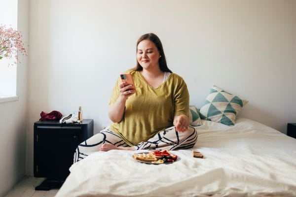 Woman sitting on bed looking at phone with breakfast on bed