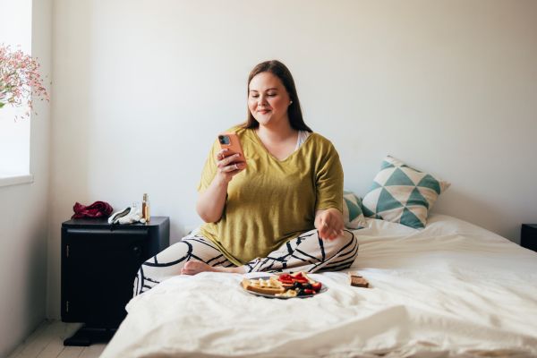 Woman sitting on bed looking at phone with breakfast on bed