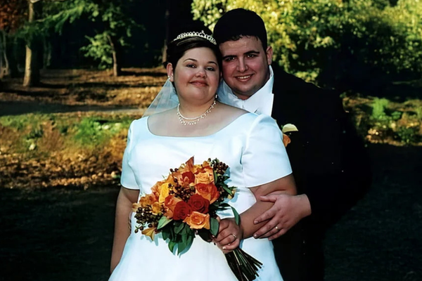 Bride and groom posing outdoors on their wedding day, with the bride in a white gown and tiara holding a bouquet of orange and yellow flowers while the groom stands behind her in a black tuxedo.