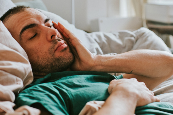 Man lying in bed with his eyes closed and hand resting on his face, illustrating symptoms that may be associated with seasonal allergies, a cold or the flu.