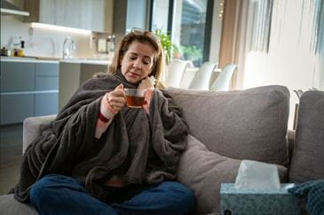 Woman sitting on the couch wrapped in a blanket, drinking hot tea and holding tissue.