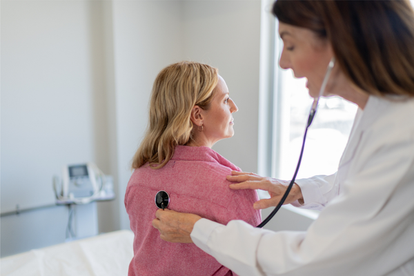 A doctor examines a woman’s breathing with a stethoscope in an exam room.