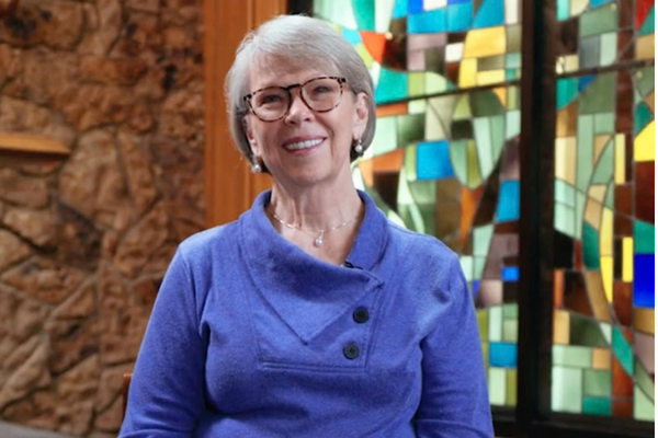 A woman with glasses smiling in a purple sweater, seated before a colorful stained-glass window.
