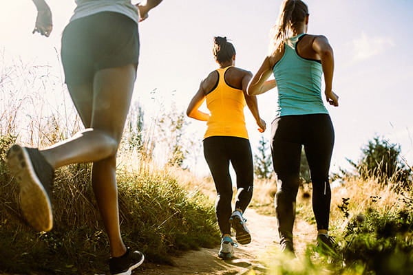 Three people jogging on a sunlit outdoor trail surrounded by grass.