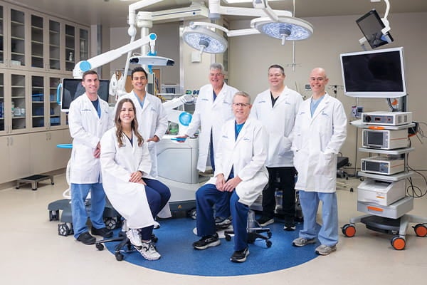 Seven orthopedic care team members in white coats pose in an operating room surrounded by surgical and imaging equipment.