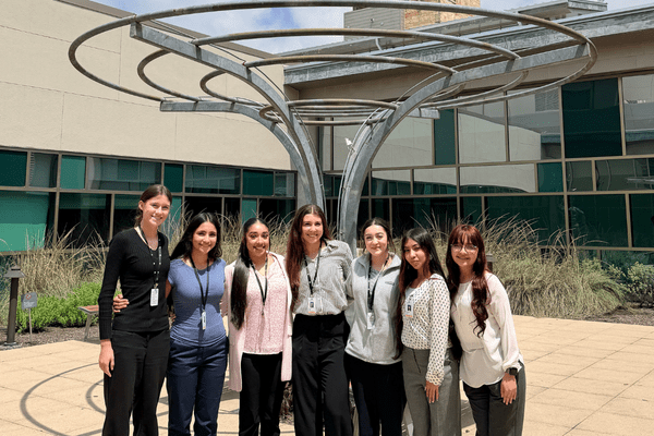 Seven student interns stand smiling together in front of Dell Children’s Medical Center.