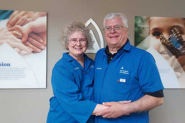 Richard and Carolyn Skurla, volunteering in blue Ascension Providence jackets, stand together in front of the hospital's Mission statement wall.