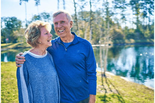 An older couple, a man and woman, practice fall prevention while walking together near a pond on a sunny day, looking healthy and smiling.