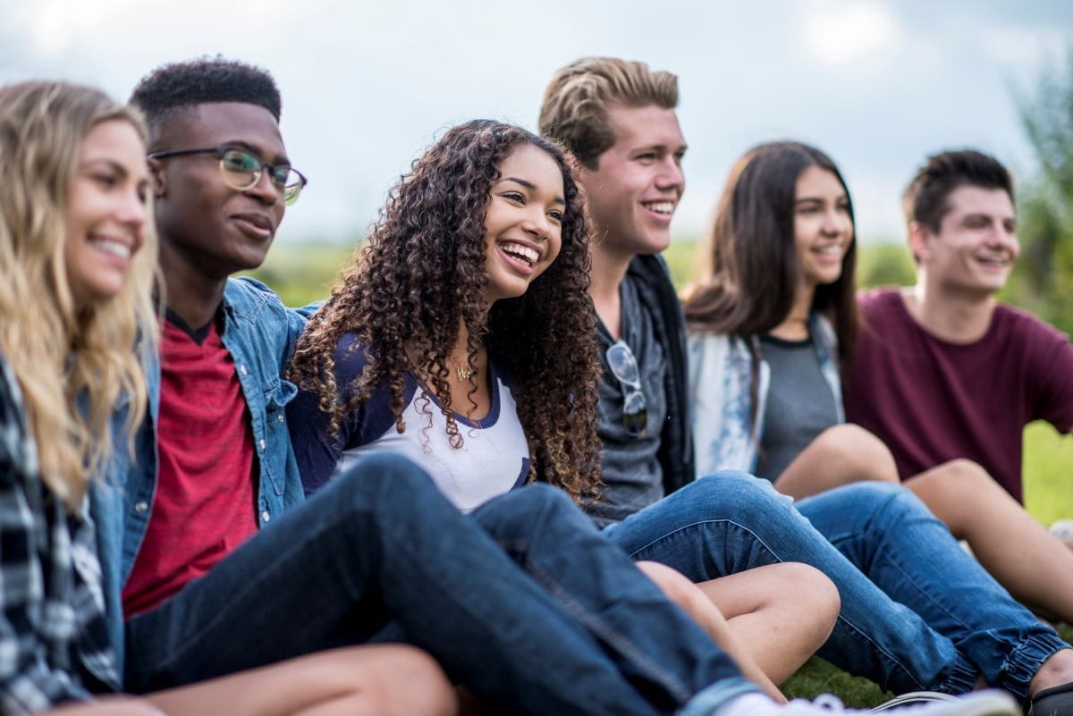 A diverse group of smiling young adults sit outdoors in a row, talking and looking toward something off camera.