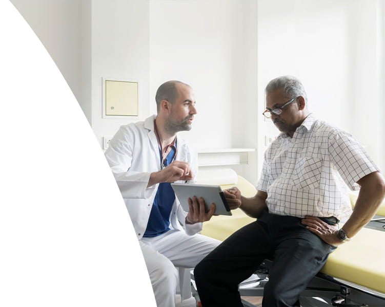 Doctor in a white coat reviewing information on a tablet with an older male patient seated on an exam table in a medical office.