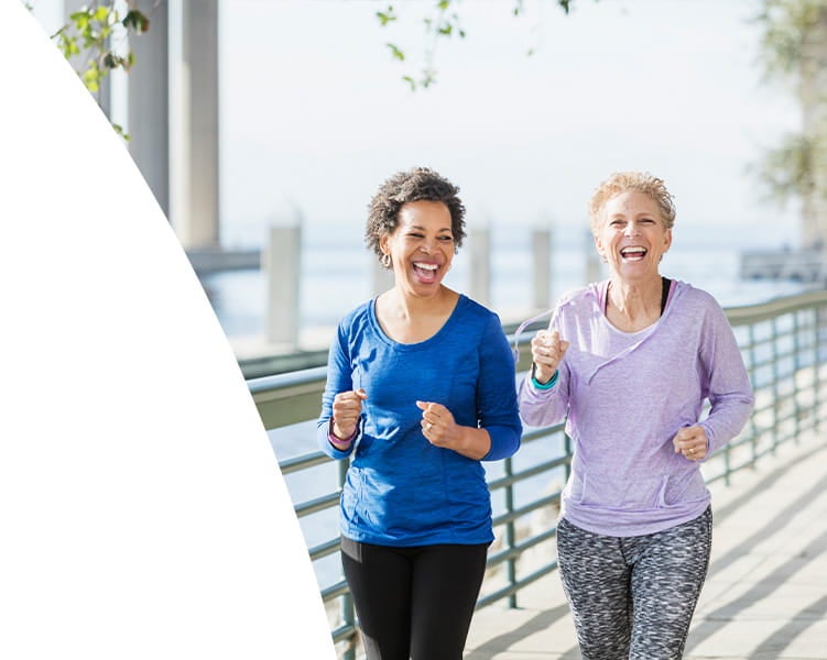 Two women jogging together on a waterfront path, smiling and holding water bottles, with a railing and cityscape in the background.