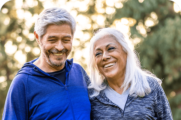Smiling older couple standing close together outdoors, with trees and warm sunlight in the background.