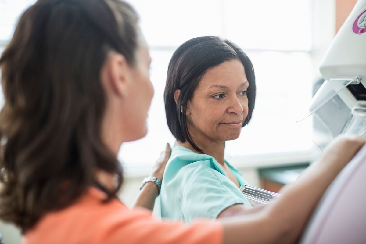 A woman receiving a mammogram from a healthcare professional, standing beside a mammography machine in a clinical setting.