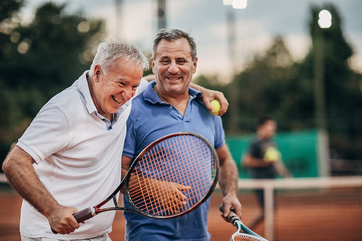 Two adult men smiling and holding tennis rackets on an outdoor tennis court during a recreational match.
