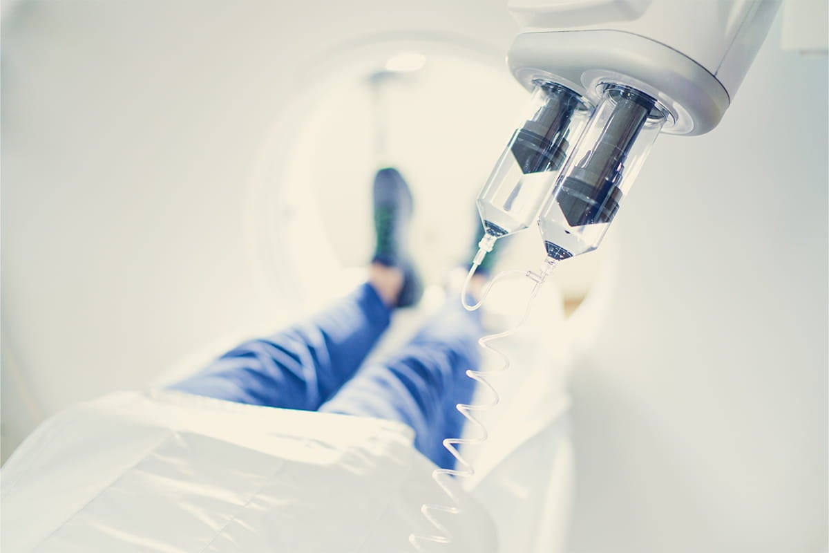 Low-angle view of a patient lying on a medical exam table with legs extended, with IV drip containers and tubing in the foreground, suggesting a clinical or hospital setting.