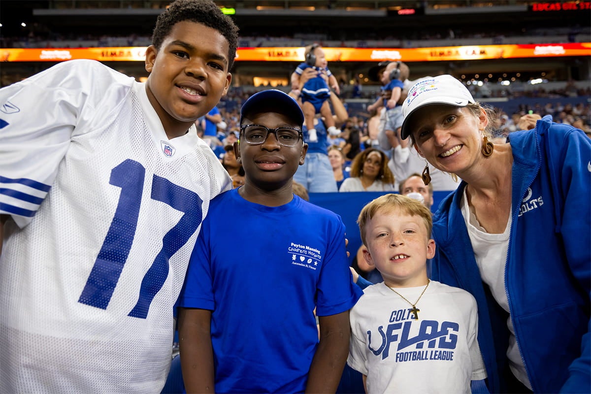 Four Indianapolis Colts fans pose together in the stadium stands, including a person wearing a number 17 jersey and children in Colts shirts