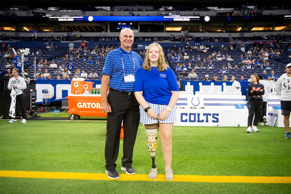 An older man and a young girl with a prosthetic leg stand together on the Indianapolis Colts football field.
